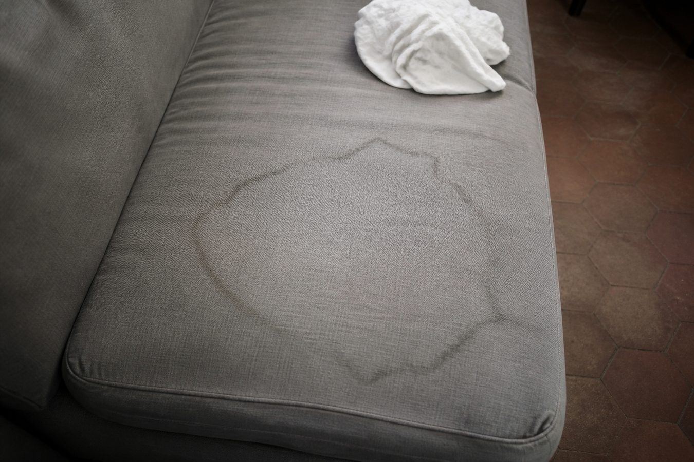 Large auréole de séchage sur l'assise d'un canapé en tissu gris, son pourtour plus foncé que le reste du tissu, un linge blanc posé à côté
